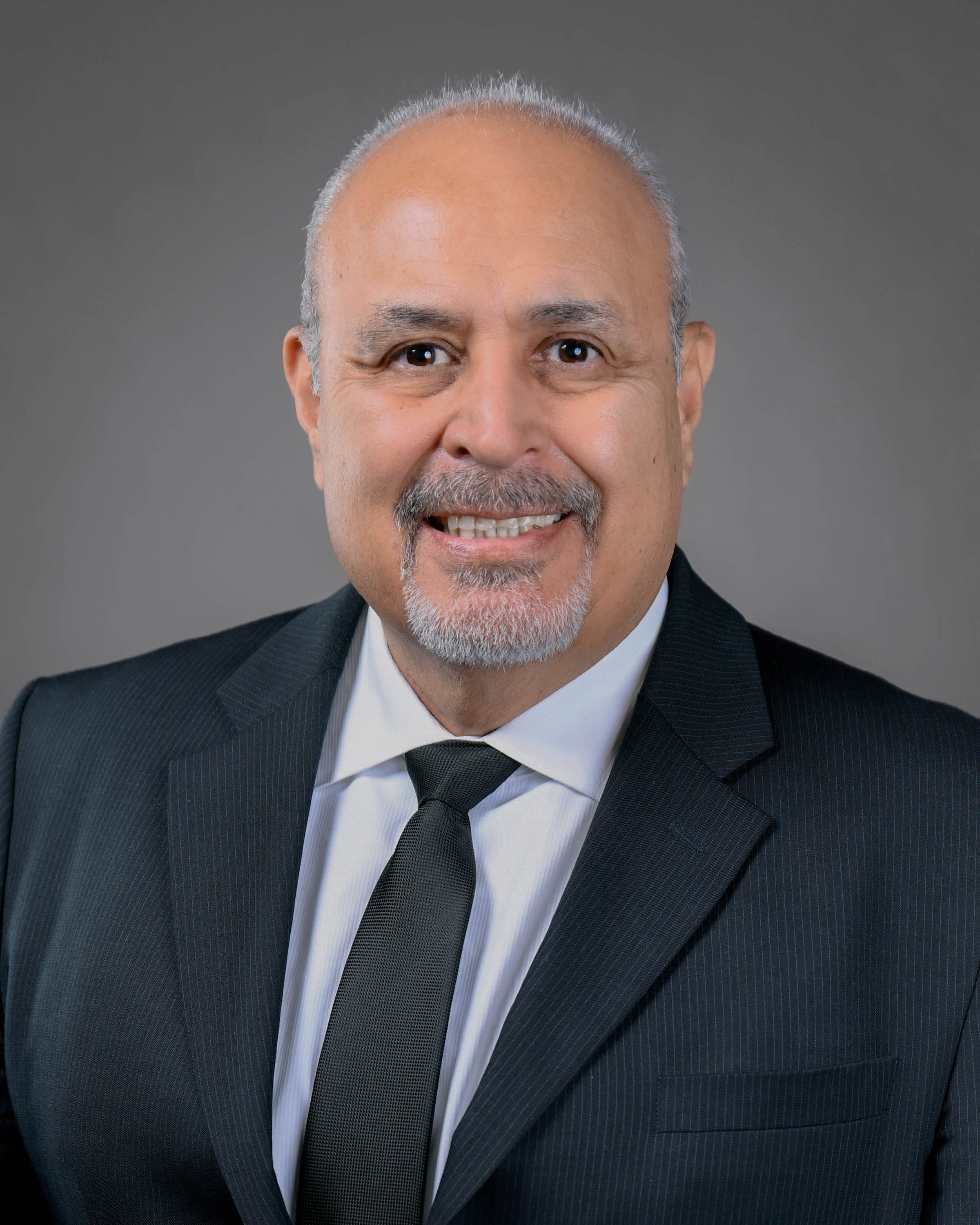 Smiling man with silver hair and goatee in a dark suit with a gray shirt and dark tie.